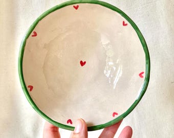 May include: A small, round ceramic bowl with a cream-coloured interior and a green rim. The bowl is decorated with small red heart shapes around the edge and one in the centre. The bowl is held by a hand.