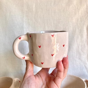 Peut inclure: Une tasse en céramique faite à la main avec une forme organique irrégulière et une finition blanc cassé brillante. L'extérieur est décoré de petits cœurs rouges peints à la main dispersés sur la surface, y compris sur l'anse incurvée.