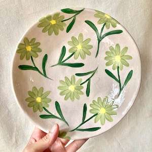 以下が含まれることがあります： ベージュの背景に、丸い陶器の皿。緑色のデイジーの花と緑色の茎で飾られています。各花の中心は黄色です。お皿は手で持たれています。