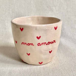May include: A small, cream-coloured ceramic cup adorned with red heart motifs and the French phrase "mon amour" in red script. The cup has a rounded form and is suitable for small items or as a decorative item.