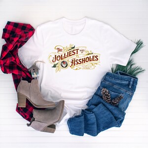 May include: White t-shirt with a gold and red graphic that says "The Jolliest Bunch of Assholes". The graphic is in a vintage style with a Santa Claus head in the center. The shirt is laid out on a white wooden surface with a red and black plaid shirt, a pair of brown boots, and a pair of blue jeans.