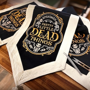 May include: Black fabric table runners with cream-colored borders and tassels. The center features gold-colored text reading SPECIMEN PRETTY LITTLE DEAD THINGS surrounded by illustrations of skeletons and ornate decorative scrollwork.