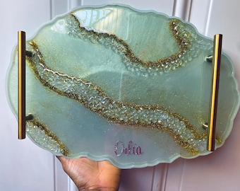 Puede incluir: Una bandeja de servir azul claro y dorada con un diseño ondulado y asas doradas. La bandeja está personalizada con el nombre "Olivia".