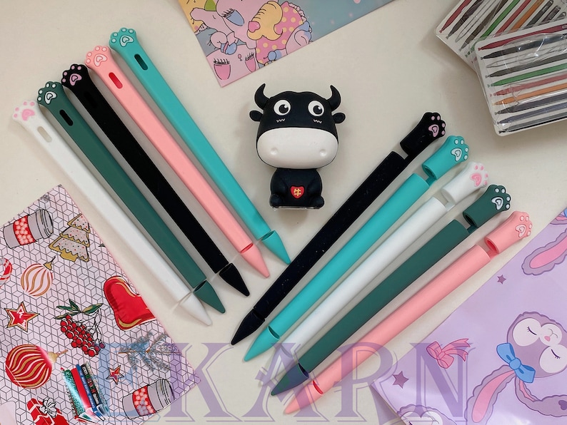 May include: Assortment of colorful silicone iPad stylus pen tips with paw print designs. The tips are arranged on a table with a small black and white bull figurine in the center.