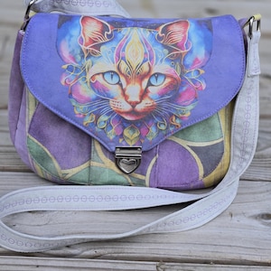 Puede incluir: Un bolso de hombro morado con una solapa con un diseño de cara de gato colorido. El bolso tiene una correa gris claro y un cierre plateado. El cuerpo del bolso es un patchwork de tela morada y verde.