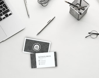 Könnte beinhalten: Zwei Visitenkarten mit einem grauen und weißen Design. Die obere Karte zeigt ein Logo mit einem Gebäude in einem Kreis. Die untere Karte zeigt den Text "Kenson Bros. Studio of Architecture" mit Kontaktinformationen darunter.