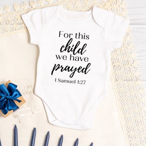 May include: White baby onesie with black text that reads "For this child we have prayed 1 Samuel 1:27".