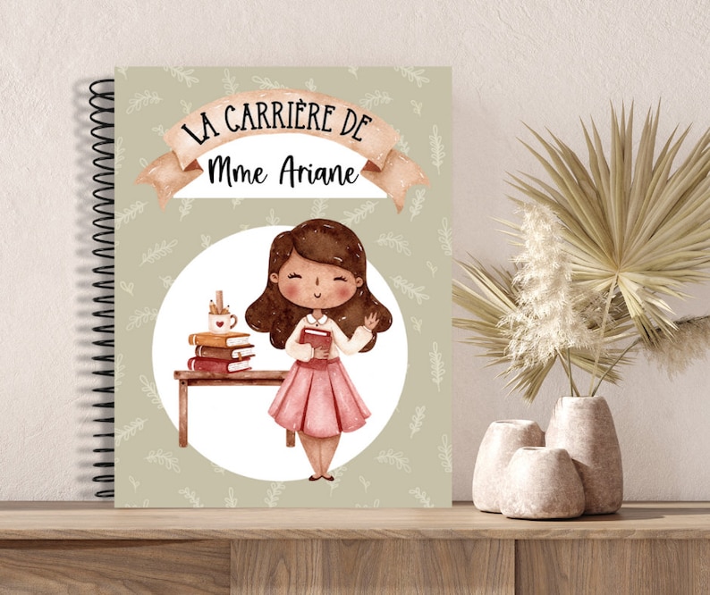 K&ouml;nnte beinhalten: Spiralgebundenes Notizbuch mit dem Text "LA CARRI&Egrave;RE DE Mme Ariane" &uuml;ber einer Illustration einer Lehrerin, die ein Buch h&auml;lt. Das Notizbuch hat einen hellgr&uuml;nen Hintergrund mit einem Bl&auml;ttermuster und einem Cartoon einer Lehrerin.