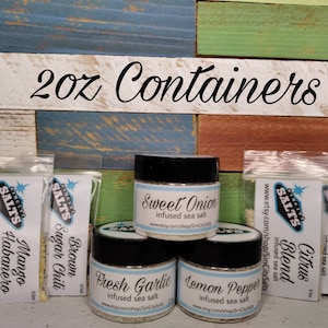 May include: Six small glass jars with lids containing various infused sea salts. The jars are labeled with the flavors: Mango Habanero, Brown Sugar Chili, Sweet Onion, Fresh Garlic, Lemon Pepper, Citrus Blend, Fresh Salsa. The jars are arranged on a wooden surface with a sign above that reads "2oz Containers".