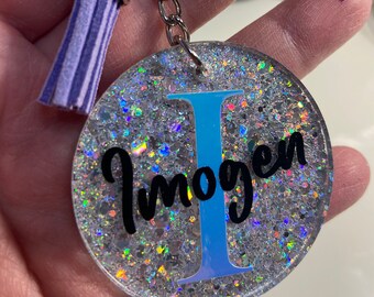 May include: A silver keyring with a purple tassel and a round, clear resin charm with iridescent glitter. The charm has a blue letter "I" and the name "Imogen" written in black.