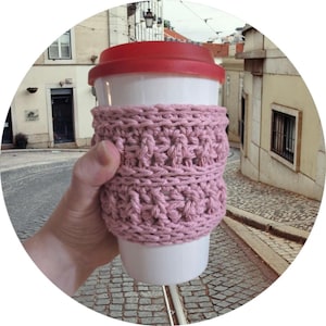 May include: A white travel mug with a red lid and a pink crocheted cozy. The cozy is wrapped around the middle of the mug, providing insulation and a decorative touch. The mug is held in a hand.