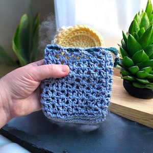 May include: A blue crocheted soap saver bag with a drawstring closure, holding a yellow and white crocheted soap. The bag is held in a hand, with a green plant in the background. The bag is made of a light blue yarn.