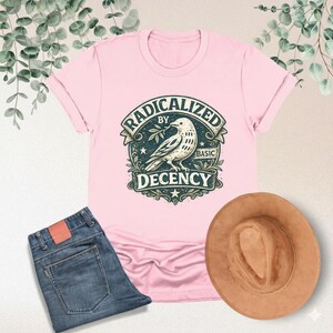 Puede incluir: Una camiseta rosa claro con el texto "RADICALIZED BY BASIC DECENCY" y un gráfico de una paloma. La imagen también incluye unos vaqueros azules y un sombrero marrón.