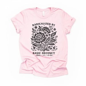 Puede incluir: Una camiseta rosa claro con cuello redondo. La camiseta presenta un diseño floral negro con el texto "RADICALIZED BY BASIC DECENCY" en una fuente estilizada. El diseño incluye flores, hojas y elementos decorativos.