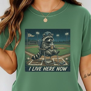 Può includere: T-shirt verde con una grafica di un procione con occhiali da sole e berretto da baseball, seduto su una sedia in un campo da baseball, che beve da una tazza. Il testo "I LIVE HERE NOW" è sotto.