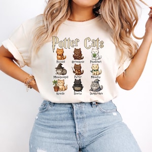May include: Cream-colored t-shirt featuring the text "Potter Cats" above illustrations of various cat characters. Each cat is depicted with unique accessories and names, such as "Rawnold" and "Meowgonagall". The shirt is worn by a person in blue jeans.