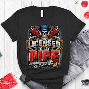 May include: Black t-shirt with a graphic featuring a skull wearing a blue cap, crossed pipe wrenches, and the text "LICENSED TO LAY PIPE" in red and gold. A wrench and pipe fittings are also shown.