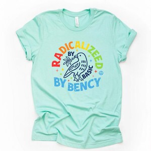 Puede incluir: Camiseta azul claro con un gráfico colorido. El gráfico presenta una paloma con una rama de olivo, rodeada por las palabras "RADICALIZED BY BASIC BY BENCY" en un arco iris. También se incluye un símbolo de la paz.
