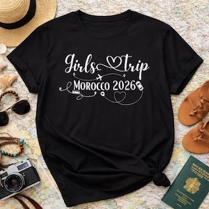 May include: Black t-shirt with the text "Girls Trip Morocco 2026" in white script. The design includes a heart, airplane, and other travel-related icons. The shirt is folded, with a map, sunglasses, and sandals in the background.