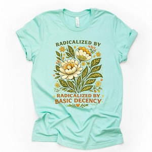 Puede incluir: Una camiseta azul claro con el texto "RADICALIZED BY BASIC DECENCY" en un diseño floral. El diseño presenta flores color crema, hojas verdes y pequeñas flores amarillas.