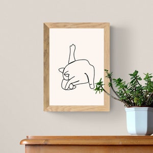 May include: A black and white line drawing of a cat stretching, framed in a light wood frame. The frame is hanging on a white wall above a wooden surface.
