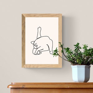 May include: A black and white line drawing of a cat stretching, framed in a light wood frame. The frame is hanging on a white wall, and a small green plant is in a white pot on a wooden surface below the frame.