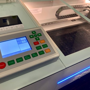 May include: A white laser cutter with a control panel featuring a digital display, buttons, and a red reset button. The laser cutter has a clear lid and a black honeycomb cutting bed.