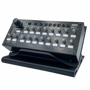 Puede incluir: Secuenciador de pasos KORG SQ-1 negro con botones y perillas blancas. El secuenciador está en un soporte negro.