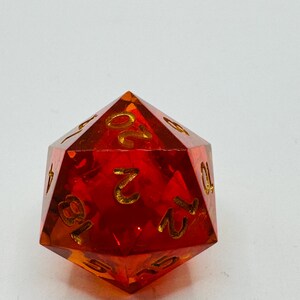 May include: A transparent, red polyhedral die with gold-colored numbers. The twenty-sided die is designed for tabletop role-playing games. The numbers are clearly visible against the red background. The die is a standard size.