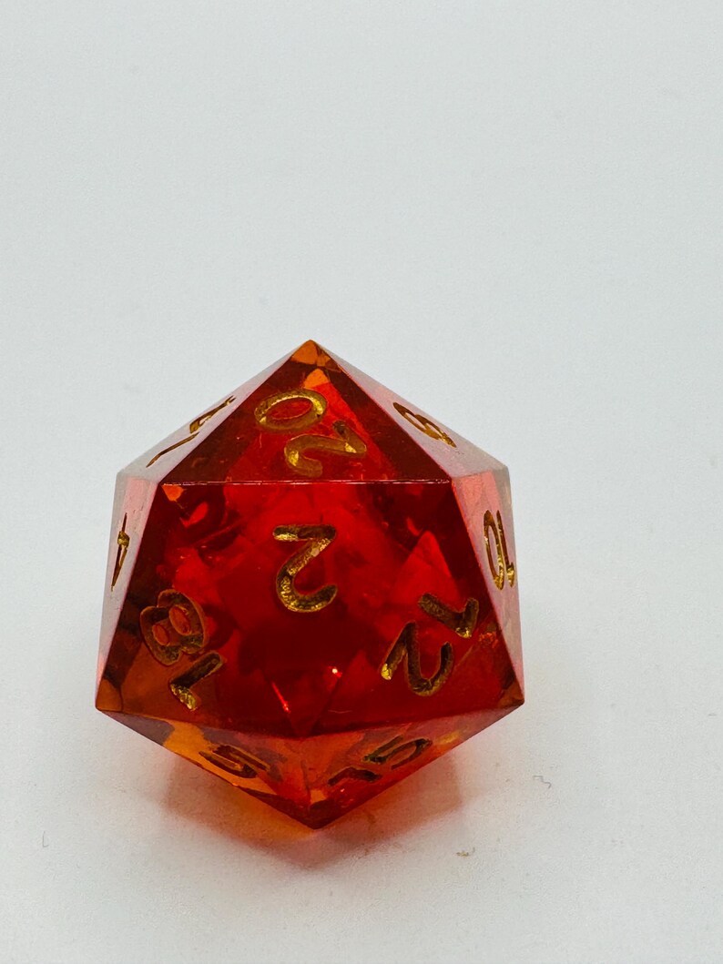 May include: A transparent, red polyhedral die with gold-colored numbers. The twenty-sided die is designed for tabletop role-playing games. The numbers are clearly visible against the red background. The die is a standard size.