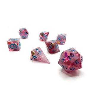 May include: A set of seven polyhedral dice in various shapes. The dice are a translucent pink with blue numbers and small candy-themed decorations inside. The dice are scattered on a white surface.