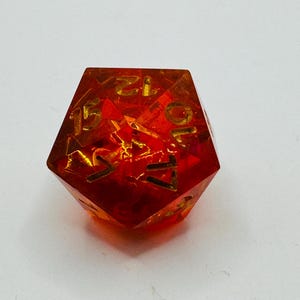 May include: A translucent, red and orange polyhedral die with gold-colored numbers. The die has a multi-faceted shape and is designed for tabletop gaming. The numbers are clearly visible against the vibrant background.