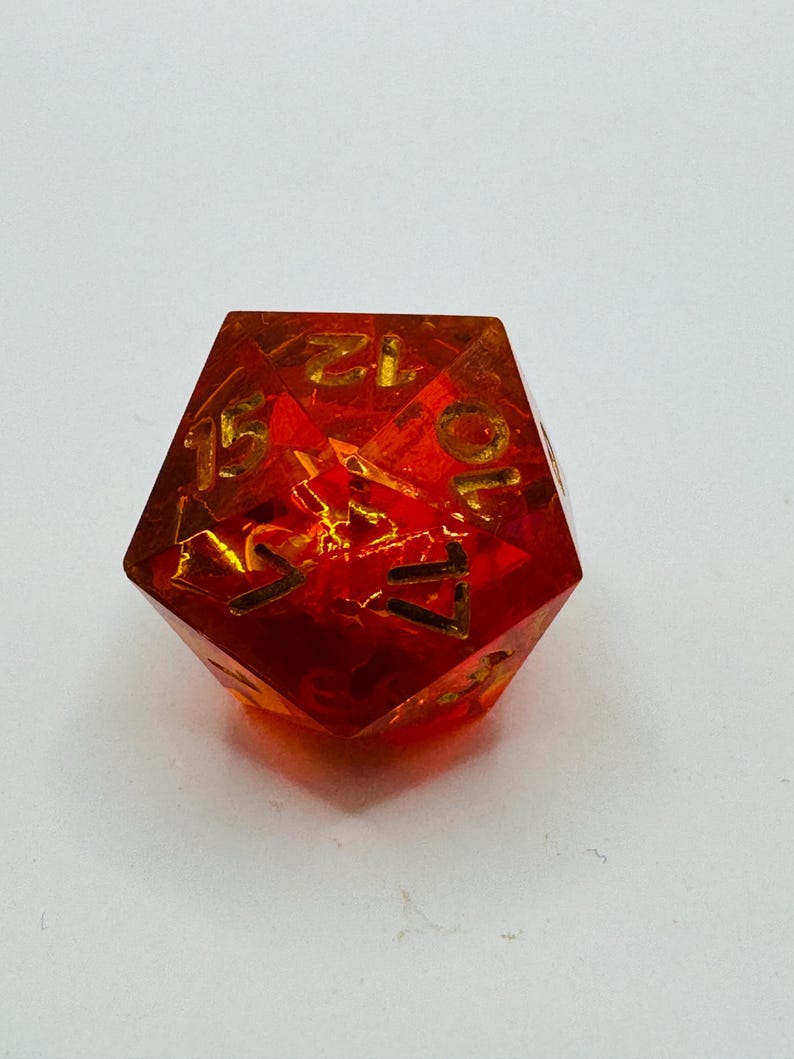 May include: A translucent, red and orange polyhedral die with gold-colored numbers. The die has a multi-faceted shape and is designed for tabletop gaming. The numbers are clearly visible against the vibrant background.