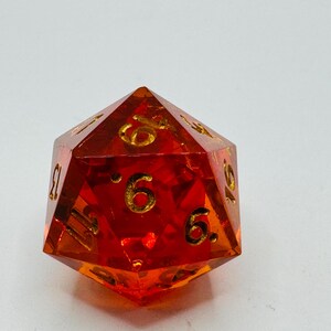 May include: A faceted, orange-red polyhedral die with gold-colored numbers. The die has 20 sides, with the numbers clearly visible. The die is made of a translucent material, allowing light to pass through. A gaming accessory.