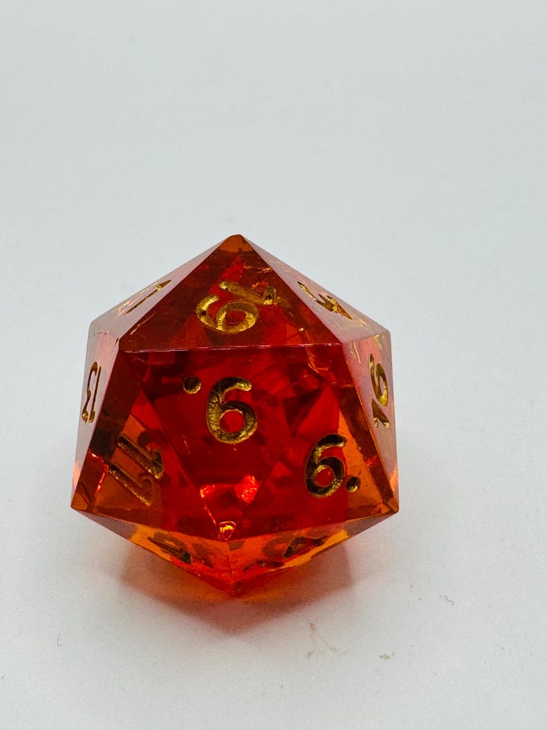 May include: A faceted, orange-red polyhedral die with gold-colored numbers. The die has 20 sides, with the numbers clearly visible. The die is made of a translucent material, allowing light to pass through. A gaming accessory.