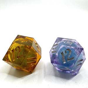 May include: Two twenty-sided dice, one amber-colored with gold glitter and one purple-colored with blue glitter. Each die features large, engraved numbers on its faces, including the number 12 prominently displayed on the front-facing side of both.