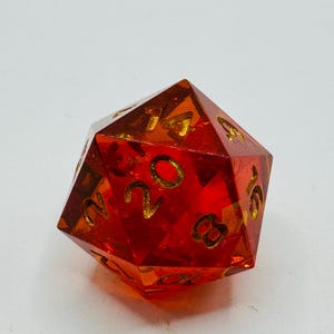 May include: A transparent, red, twenty-sided die with gold-colored numbers. The die is a polyhedron, a geometric shape with multiple flat faces. This gaming accessory is used in tabletop role-playing games.