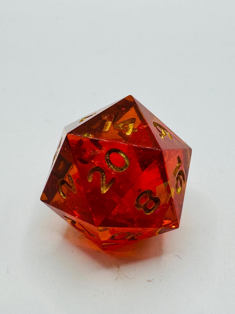 May include: A transparent, red, twenty-sided die with gold-colored numbers. The die is a polyhedron, a geometric shape with multiple flat faces. This gaming accessory is used in tabletop role-playing games.
