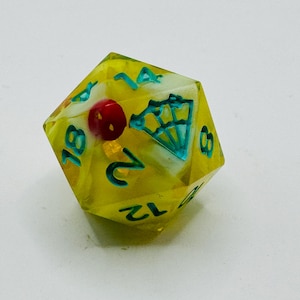 May include: A yellow, multi-sided die with teal numbers and a diamond graphic. The die has a red sphere and white accents inside. The numbers are teal and the die is made of a clear material.