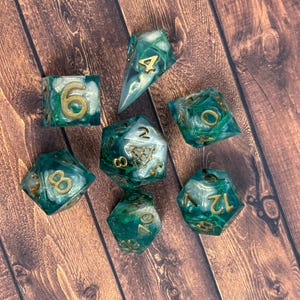 May include: A set of seven polyhedral dice with a marbled teal and white design, accented with gold numbering. The dice are arranged on a wooden surface, showcasing their unique color and design.