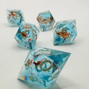 Könnte beinhalten: Ein Set aus fünf durchscheinenden, hellblauen polyedrischen Würfeln mit scharfen Kanten. Jeder Würfel enthält schwebende orangefarbene und weiße Einschlüsse, feine Glitzerpartikel und große, helle Ziffern, die in die Harzoberflächen eingraviert sind.