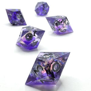 May include: A set of five translucent purple polyhedral dice with sharp edges and geometric shapes. Each die contains dark, wispy inclusions suspended within the resin, and the engraved numbers are filled with a dark, contrasting ink color.