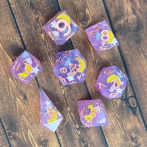 Puede incluir: Juego de siete dados de resina morada con números dorados. Cada dado presenta un diseño celestial con lunas, estrellas y nubes en amarillo y rosa. Los dados están dispuestos sobre una superficie de madera.