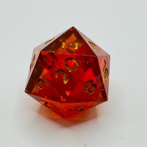 May include: A transparent, red twenty-sided die with gold-colored numbers. The die is a polyhedron, with each face displaying a number from 1 to 20. The die is designed for tabletop role-playing games.
