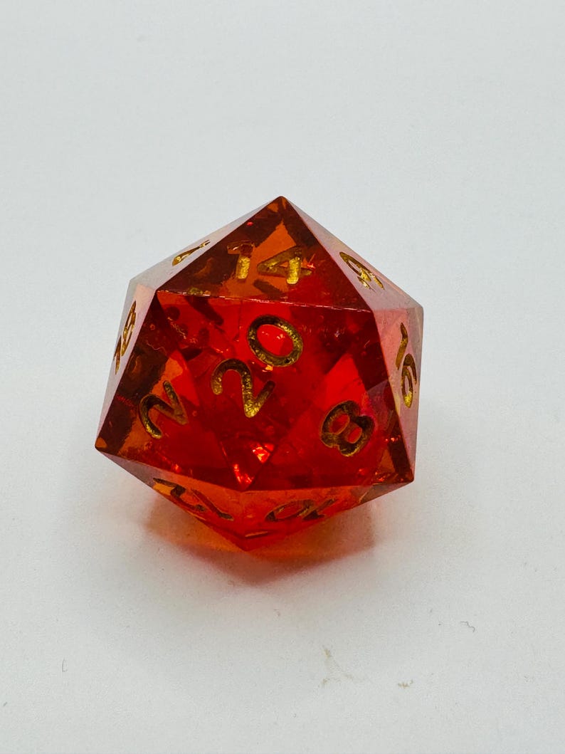 May include: A transparent, red twenty-sided die with gold-colored numbers. The die is a polyhedron, with each face displaying a number from 1 to 20. The die is designed for tabletop role-playing games.
