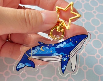 May include: A blue whale keychain with a starry night sky design. The whale is surrounded by clouds and stars. The keychain has a gold star charm attached to it.