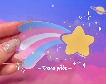 May include: A translucent sticker with a rainbow and a yellow star. The rainbow is in the colors of the transgender pride flag. The sticker has the text "_trans pride_" and "Hattiebee Designs" on it.