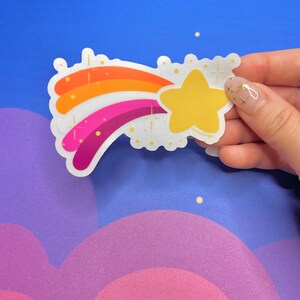 May include: A rainbow-colored shooting star sticker with a yellow star at the end. The sticker has a white background with gold glitter accents.
