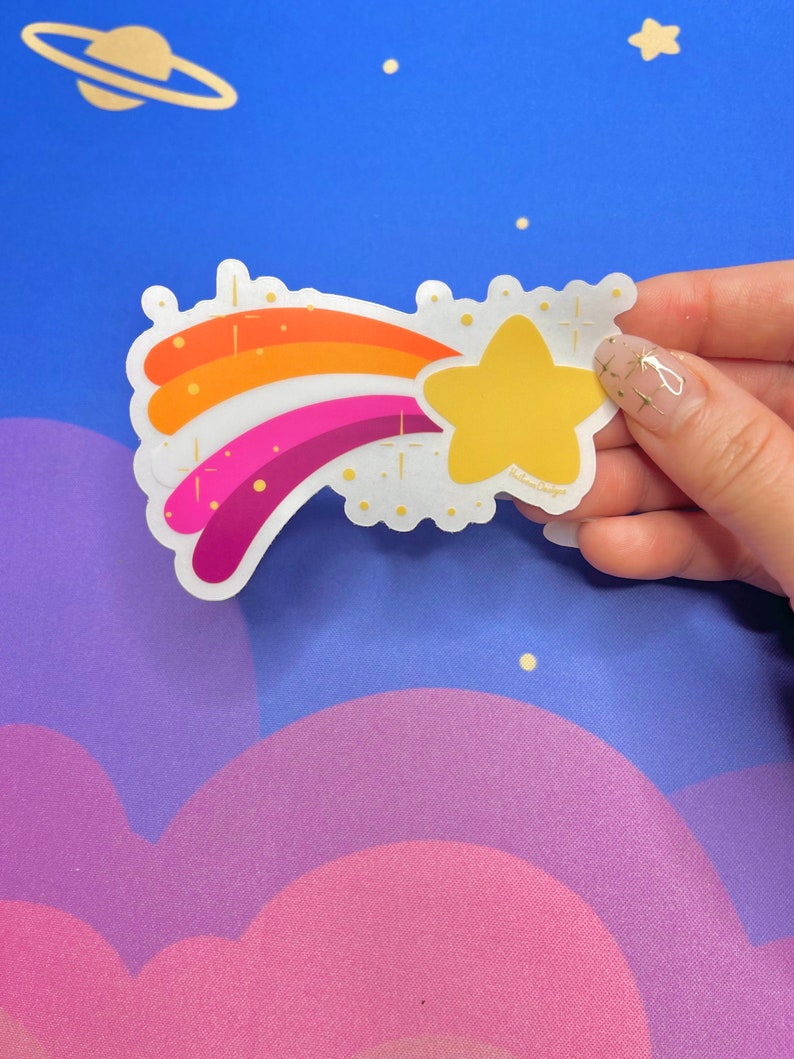 May include: A rainbow-colored shooting star sticker with a yellow star at the end. The sticker has a white background with gold glitter accents.