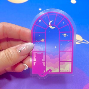May include: A clear sticker with a pink silhouette of a cat looking out a window with a crescent moon and stars in the sky. The sticker has the text "Hallowee Designs" at the bottom.